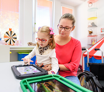 A child with glasses sits on her mother's lap while the daughter is using her tablet