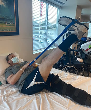 Photo of Kevin laying in bed while stretching on of his hamstrings with a stretching band