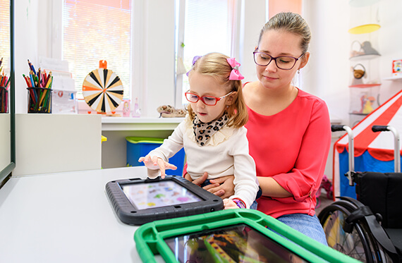 A child with glasses sits on her mother's lap while the daughter is using her tablet